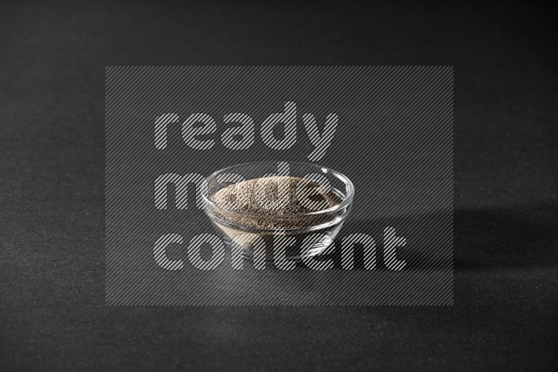 A glass bowl full of black pepper powder on black flooring