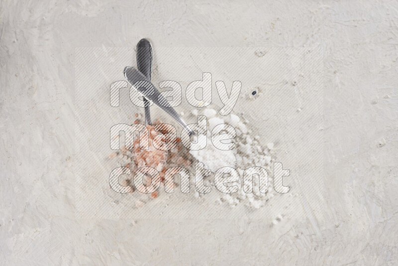 2 metal spoons filled with white salt and pink himalayan salt on white background