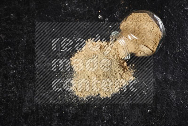 A glass jar full of ground ginger powder flipped with some spilling powder on black background