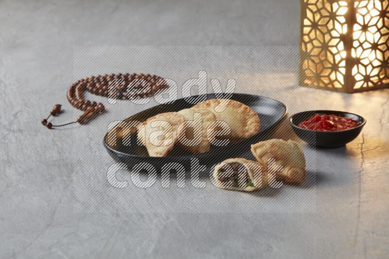 Three fried sambosas in an oval shaped black plate and a red sauce in a black round ramekin with a brown misbaha and a golden lantern on the side on a gray background