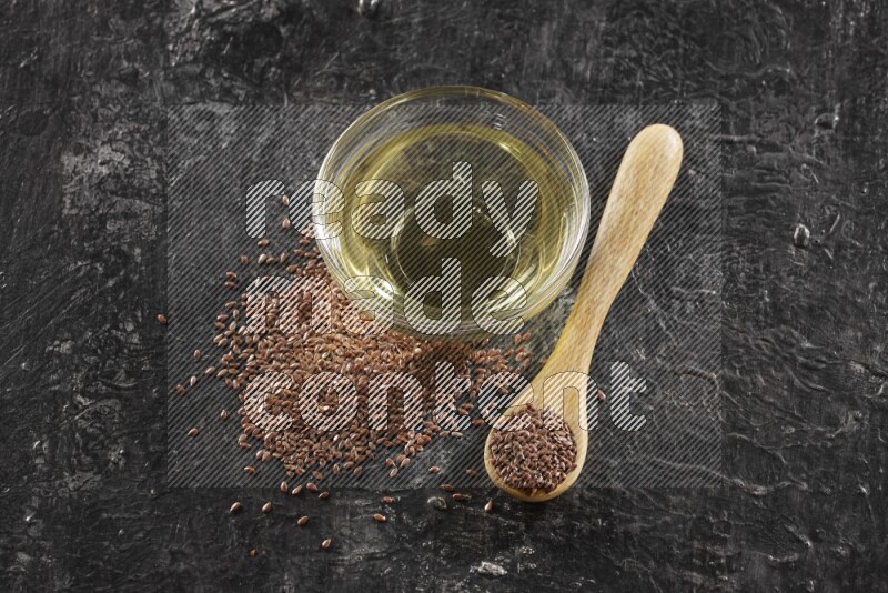 A glass bowl full of flaxseeds oil and wooden spoon full of flaxseeds with seeds spread on a textured black flooring
