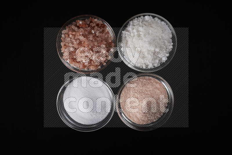 4 glass bowls filled with table salt, coarse sea salt, fine himalayan salt and coarse himalayan salt on black background