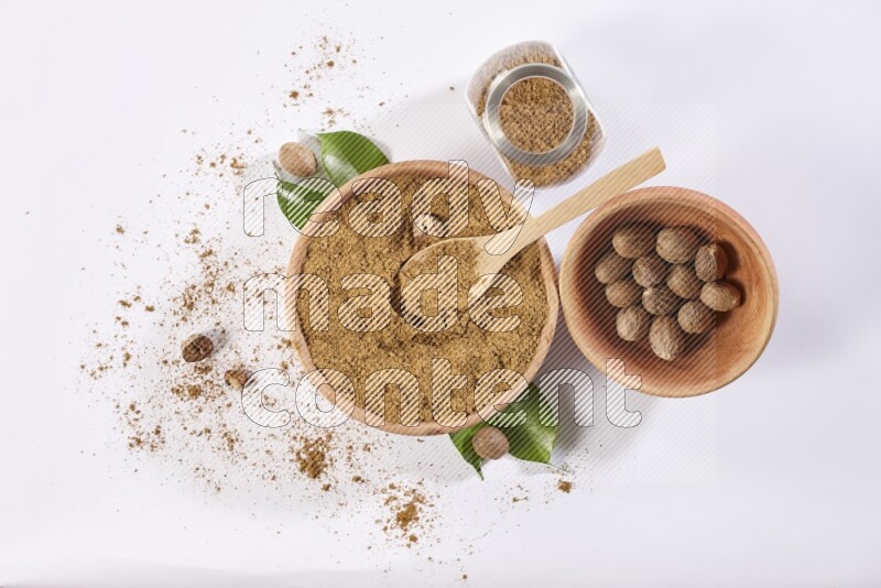 A wooden bowl with a spoon in it and glass spice jar all, full of nutmeg powder and a wooden bowl full of whole nutmeg seeds on a white flooring