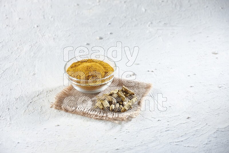 A glass bowl full of turmeric powder and dried turmeric whole finger on a piece of burlap on a textured white flooring
