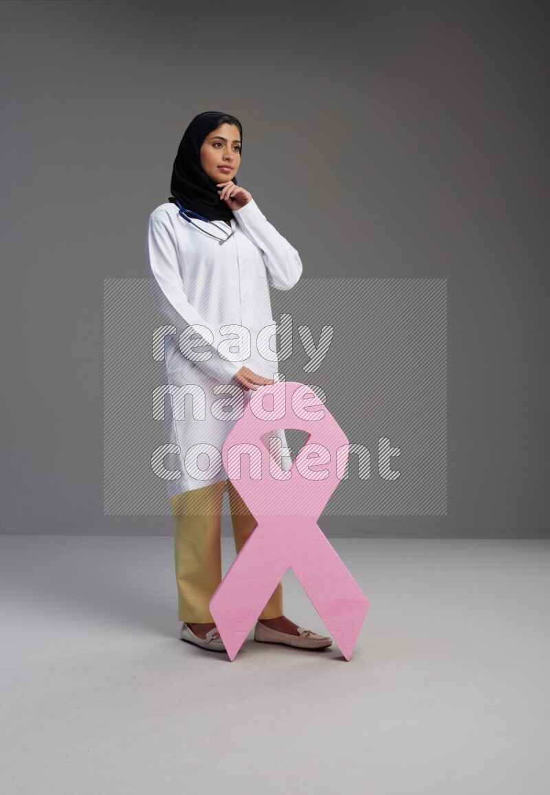 Saudi woman wearing lab coat with stethoscope standing holding awareness ribbon symbol standing on Gray background