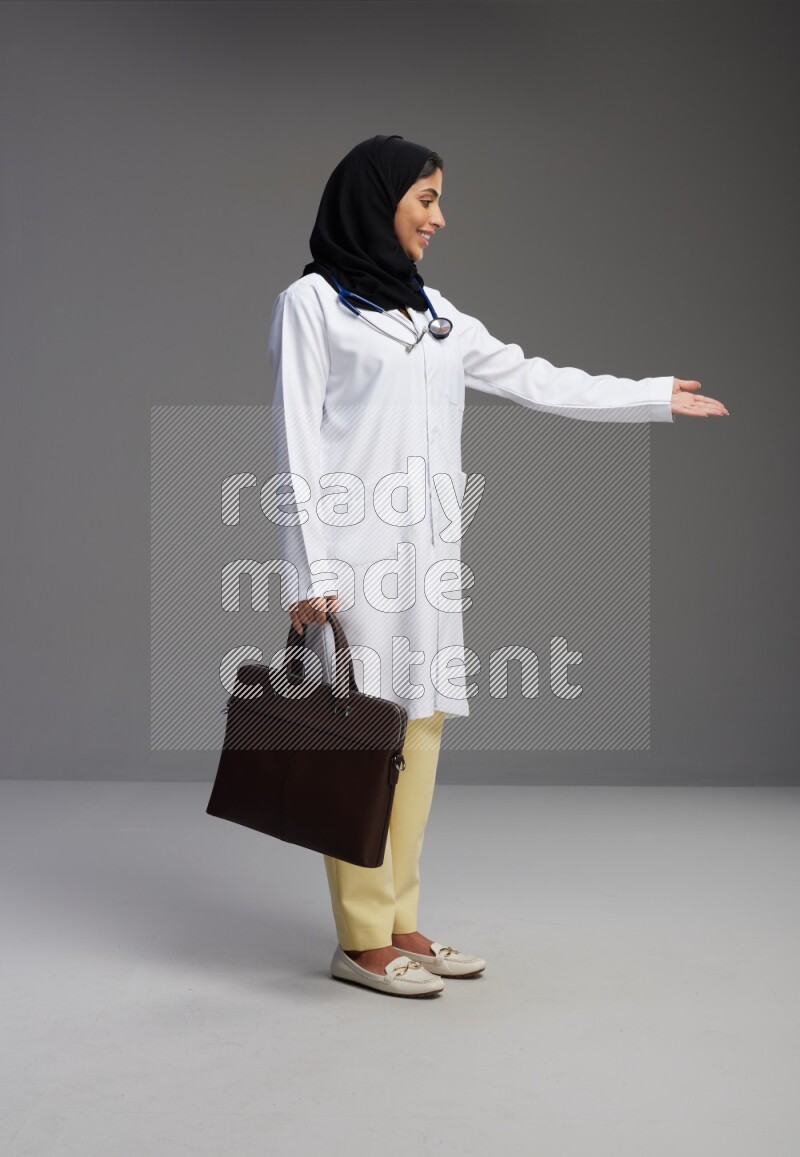Saudi woman wearing lab coat with stethoscope standing holding bag on Gray background