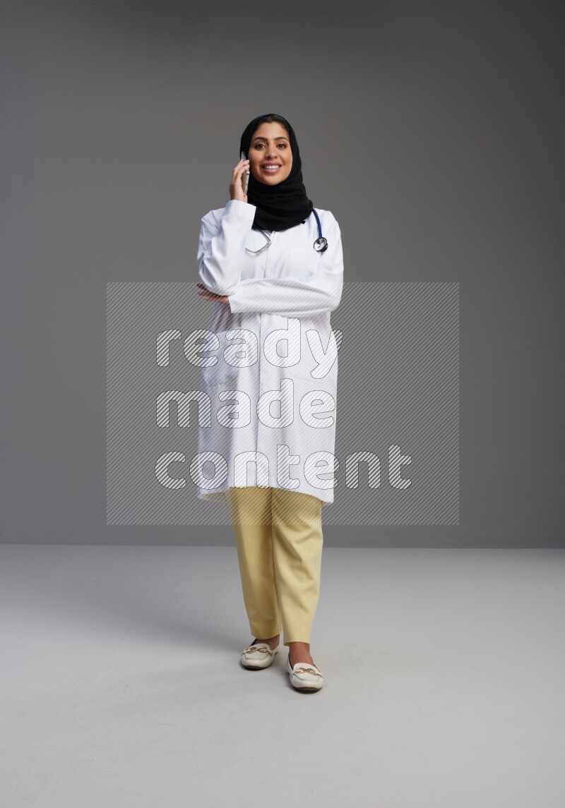 Saudi woman wearing lab coat with stethoscope standing talking on phone on Gray background