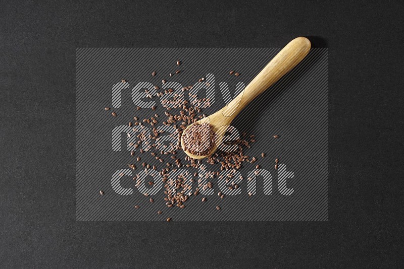 A wooden spoon full of flaxseeds and seeds spread beside it on a black flooring