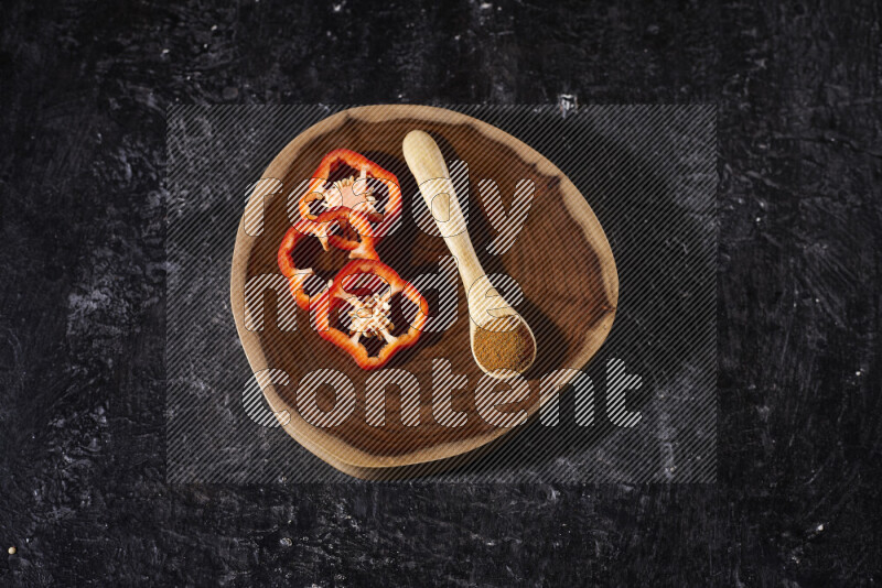 A wooden spoon full of ground paprika powder with red bell pepper slices beside it, all on a wooden tray on black background