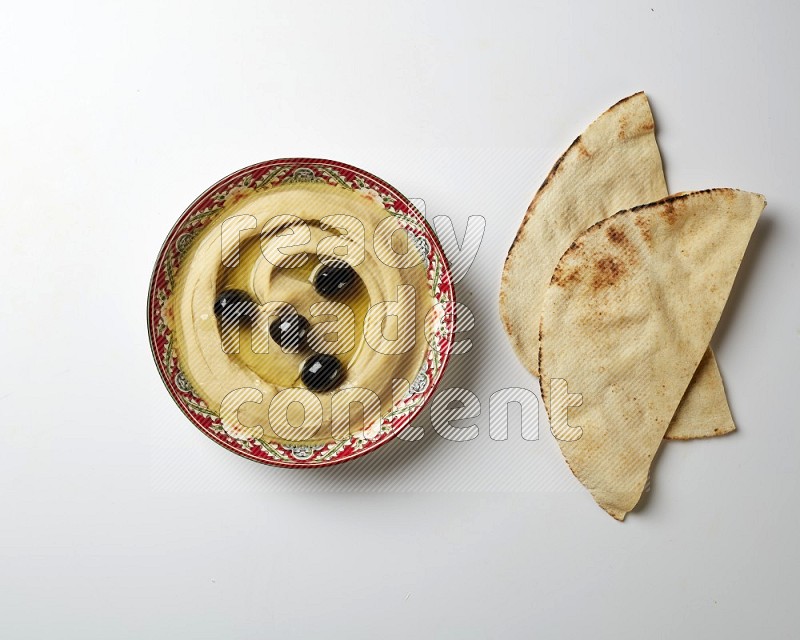 Hummus in a red plate with patterns garnished with black olives on a white background