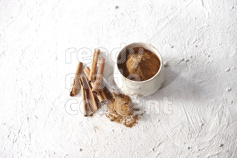 Ceramic beige bowl full of cinnamon powder and a metal spoon with cinnamon sticks next of it on a textured white background