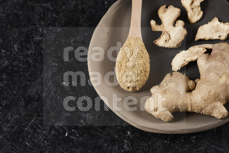 A wooden spoon full of ground ginger powder with fresh and dried ginger, all on a pottery plate on black background