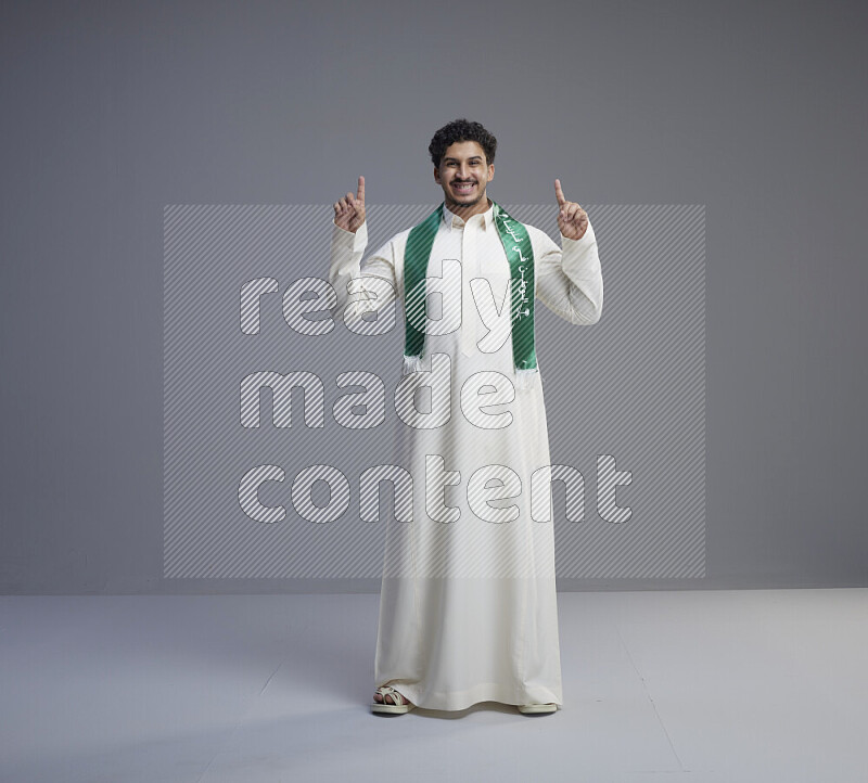 A Saudi man standing wearing thob and Saudi flag scarf on gray background