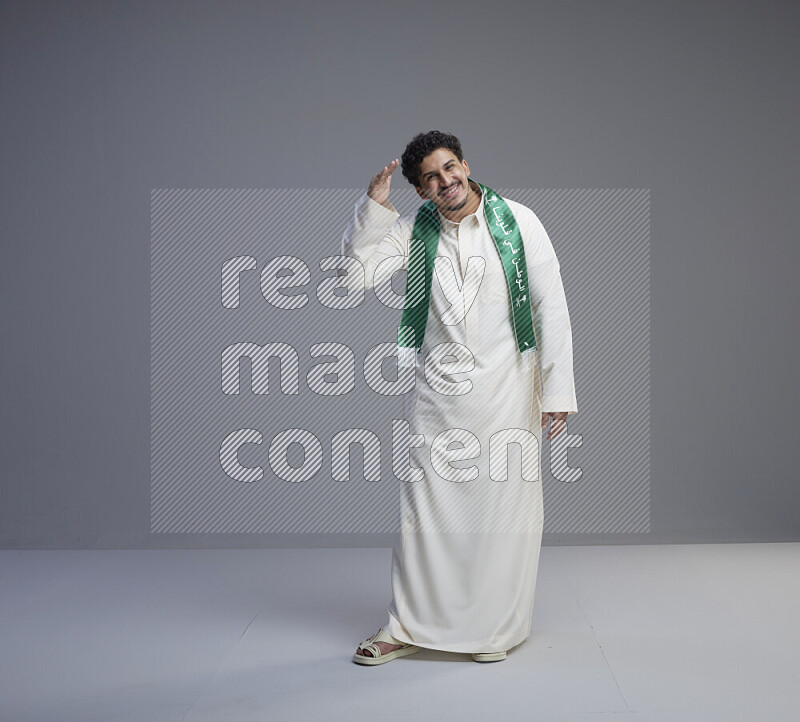 A Saudi man standing wearing thob and Saudi flag scarf on gray background