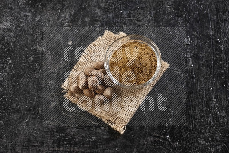 A glass bowl full of nutmeg powder with the seeds beside it on burlap fabric on a textured black flooring in different angles