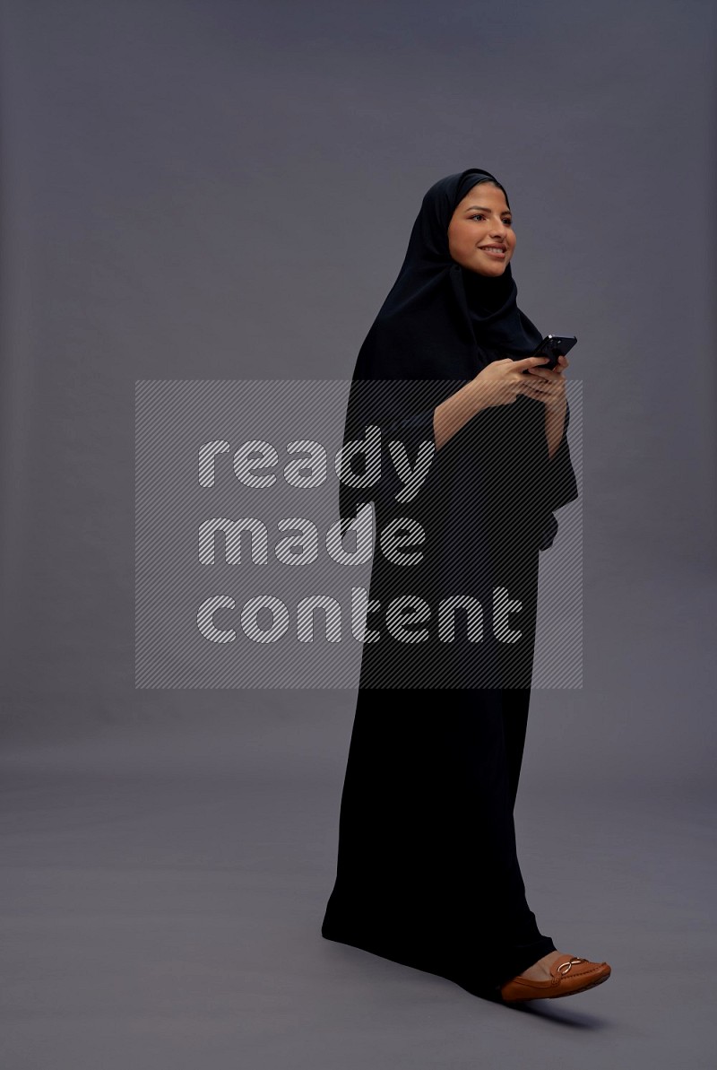 Saudi woman wearing Abaya standing texting on phone on gray background