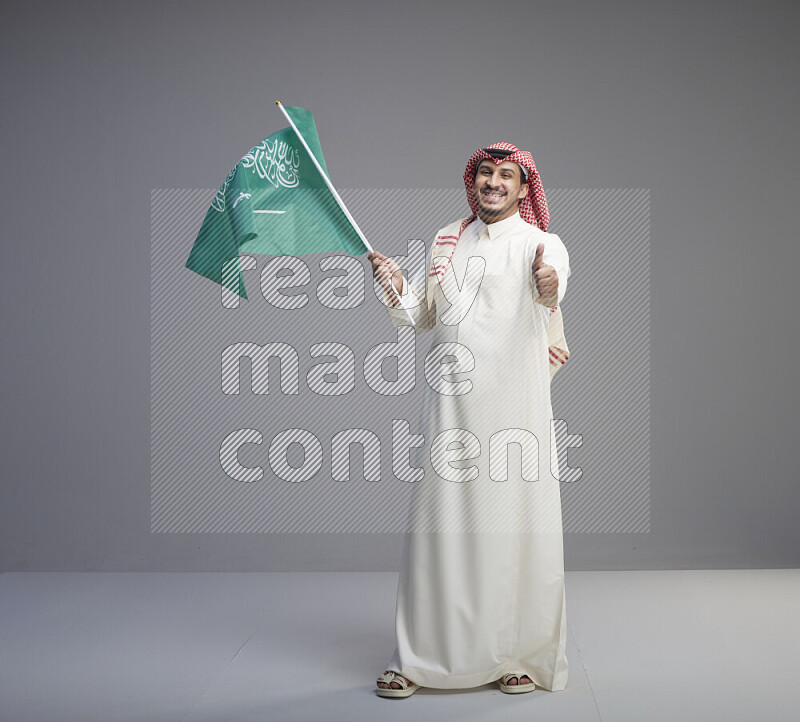 A Saudi man standing wearing thob and red shomag raising big Saudi flag on gray background