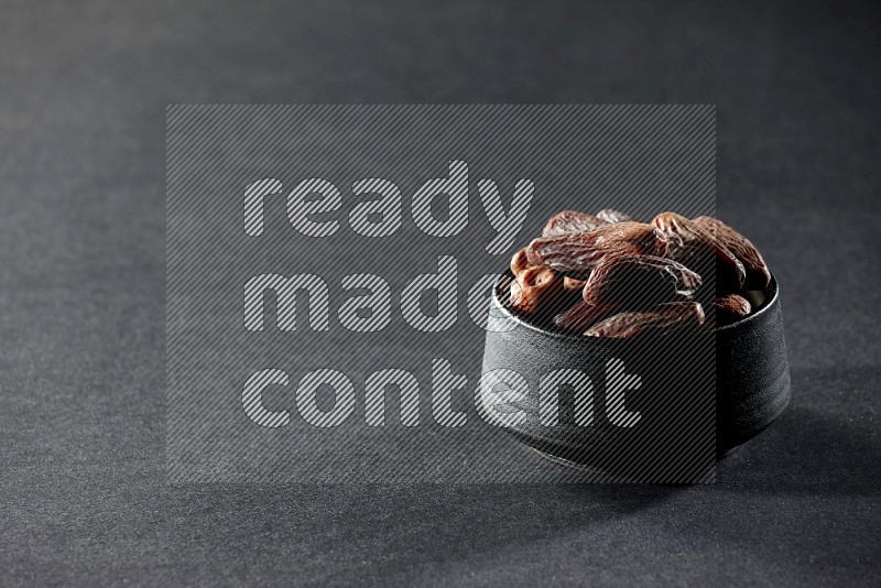 A black pottery bowl full of dried dates on a black background in different angles