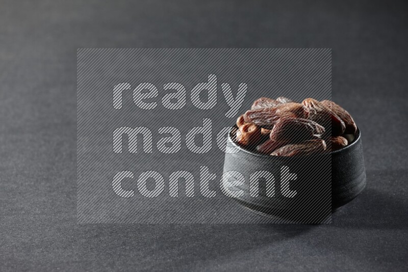 A black pottery bowl full of dried dates on a black background in different angles