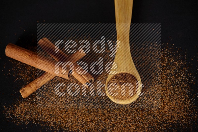 Cinnamon powder in a wooden spoon and cinnamon sticks beside it on black background