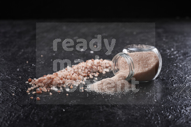 A glass jar full of fine himalayan salt with some himalayan crystals beside it on a black background