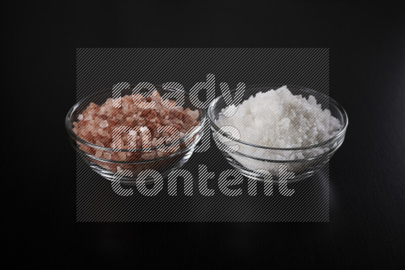 2 glass bowls one is filled with himalayan salt and the other with white salt on black background