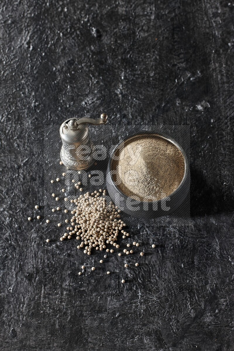 Black pottery bowl full of white pepper powder with pepper beads and metal pepper grinder on textured black flooring