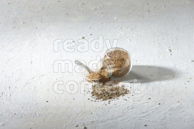A glass spice jar and metal spoon full of cumin powder and the jar flipped and powder spilled out with cumin seeds on textured white flooring