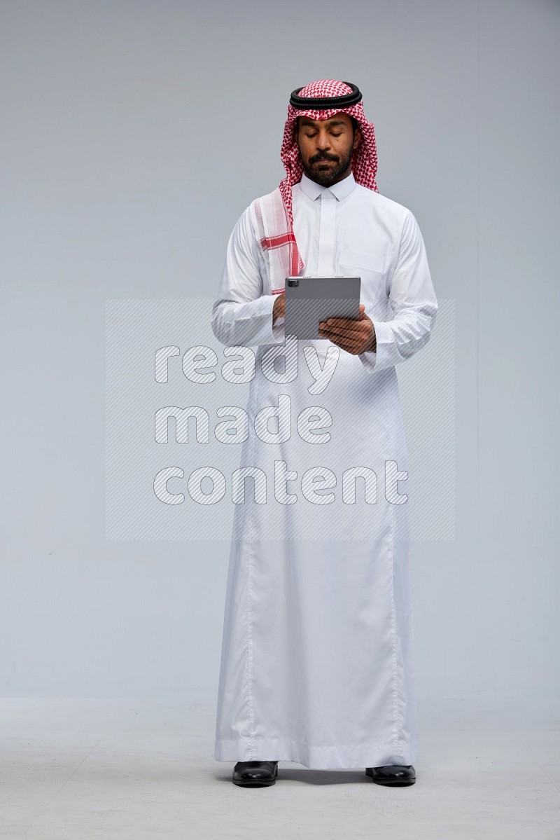 Saudi man Wearing Thob and shomag standing working on tablet on Gray background