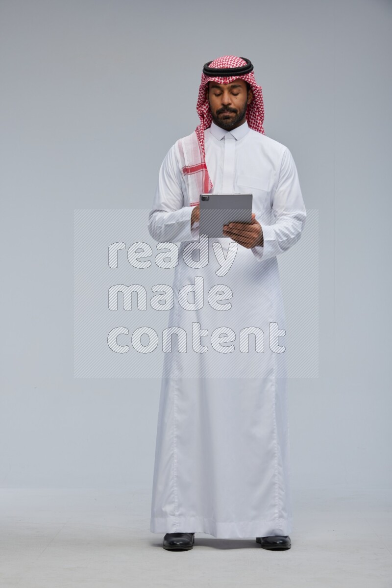 Saudi man Wearing Thob and shomag standing working on tablet on Gray background