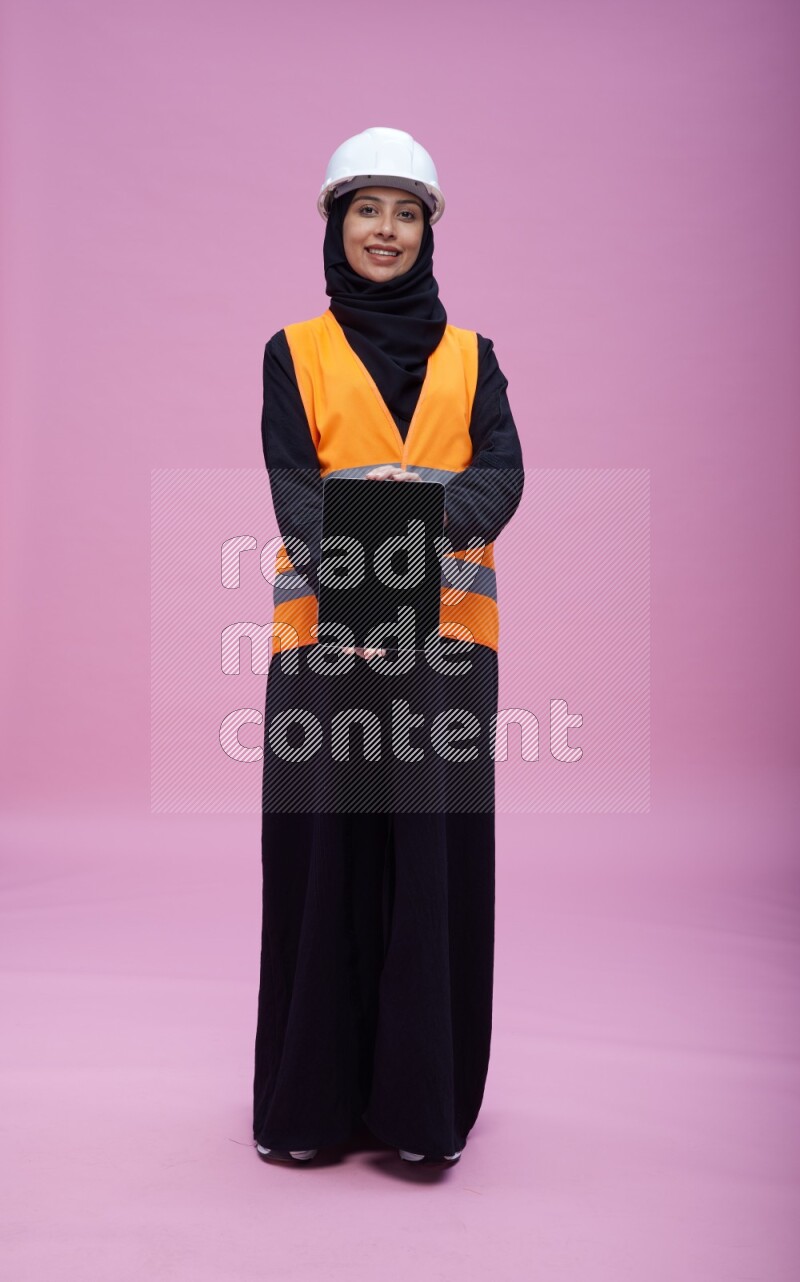 Saudi woman wearing Abaya with engineer vest and helmet standing showing tablet to camera on pink background