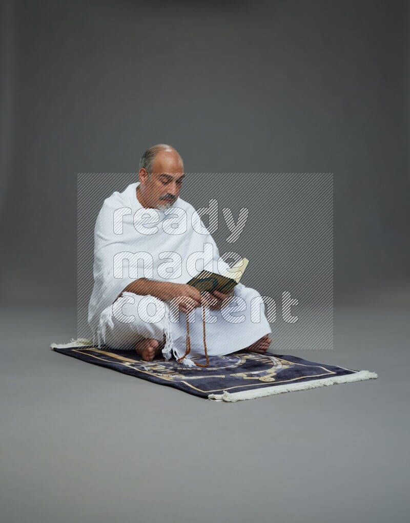 A man wearing Ehram sitting on mate prayer reading quran on gray background