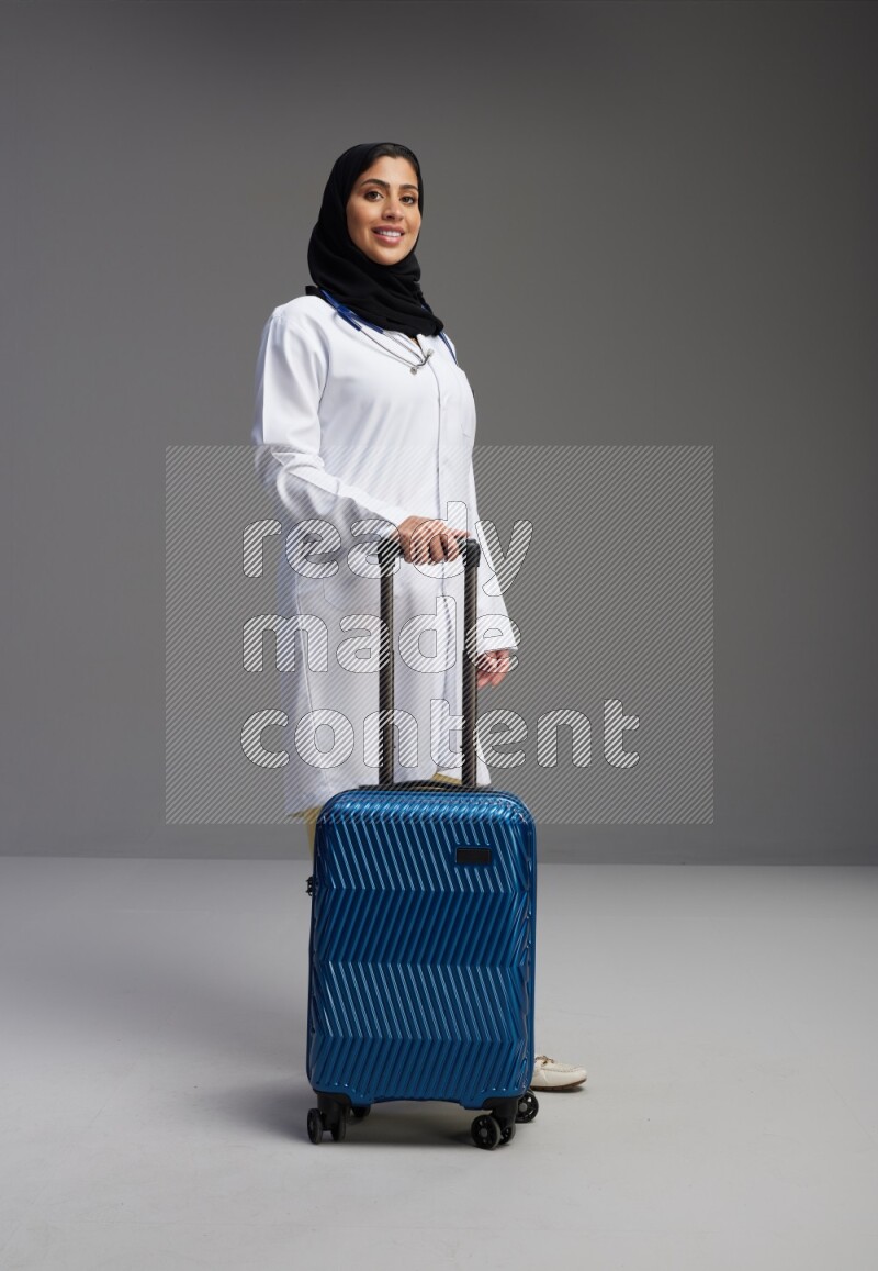 Saudi woman wearing lab coat with stethoscope standing holding Travel bag on Gray background