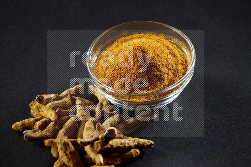 A glass bowl full of turmeric powder and dried whole fingers beside it on black flooring