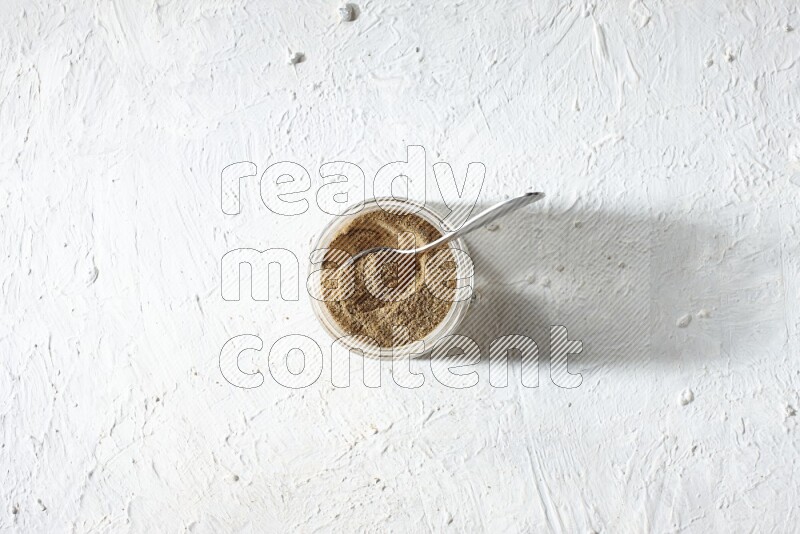 A glass jar and a metal spoon full of cumin powder on textured white flooring