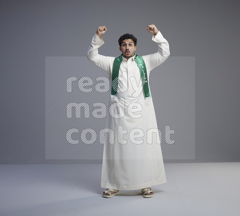 A Saudi man standing wearing thob and Saudi flag scarf on gray background