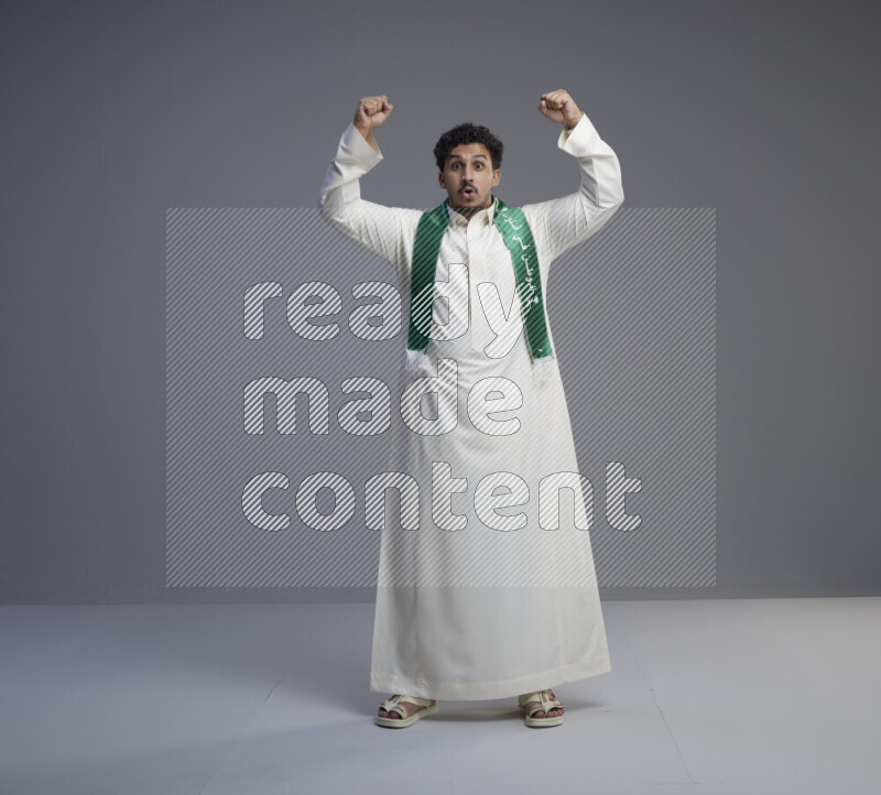 A Saudi man standing wearing thob and Saudi flag scarf on gray background
