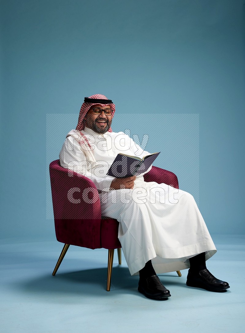 Saudi Man with shimag sitting on chair reading book on blue background