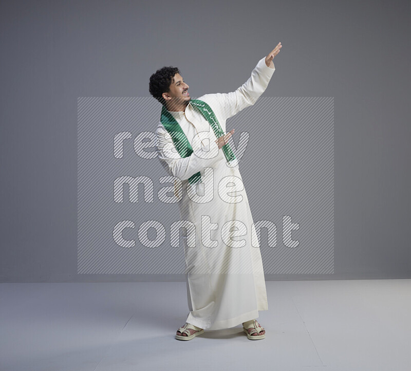 A Saudi man standing wearing thob and Saudi flag scarf on gray background