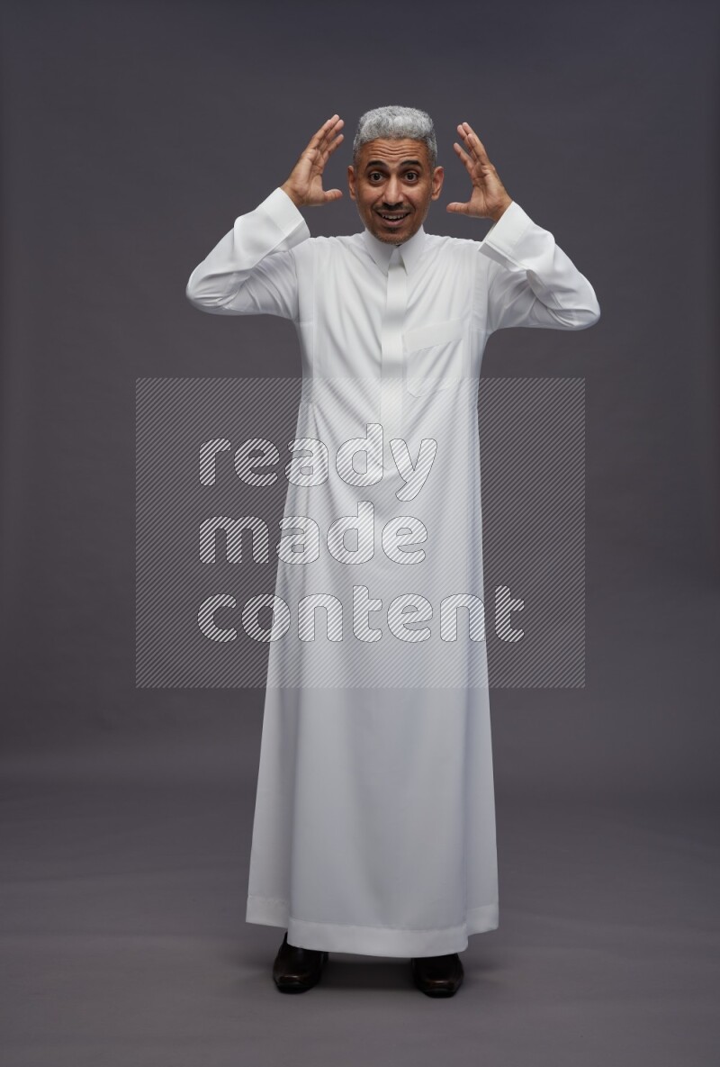 Saudi man wearing thob standing hands behind head on gray background