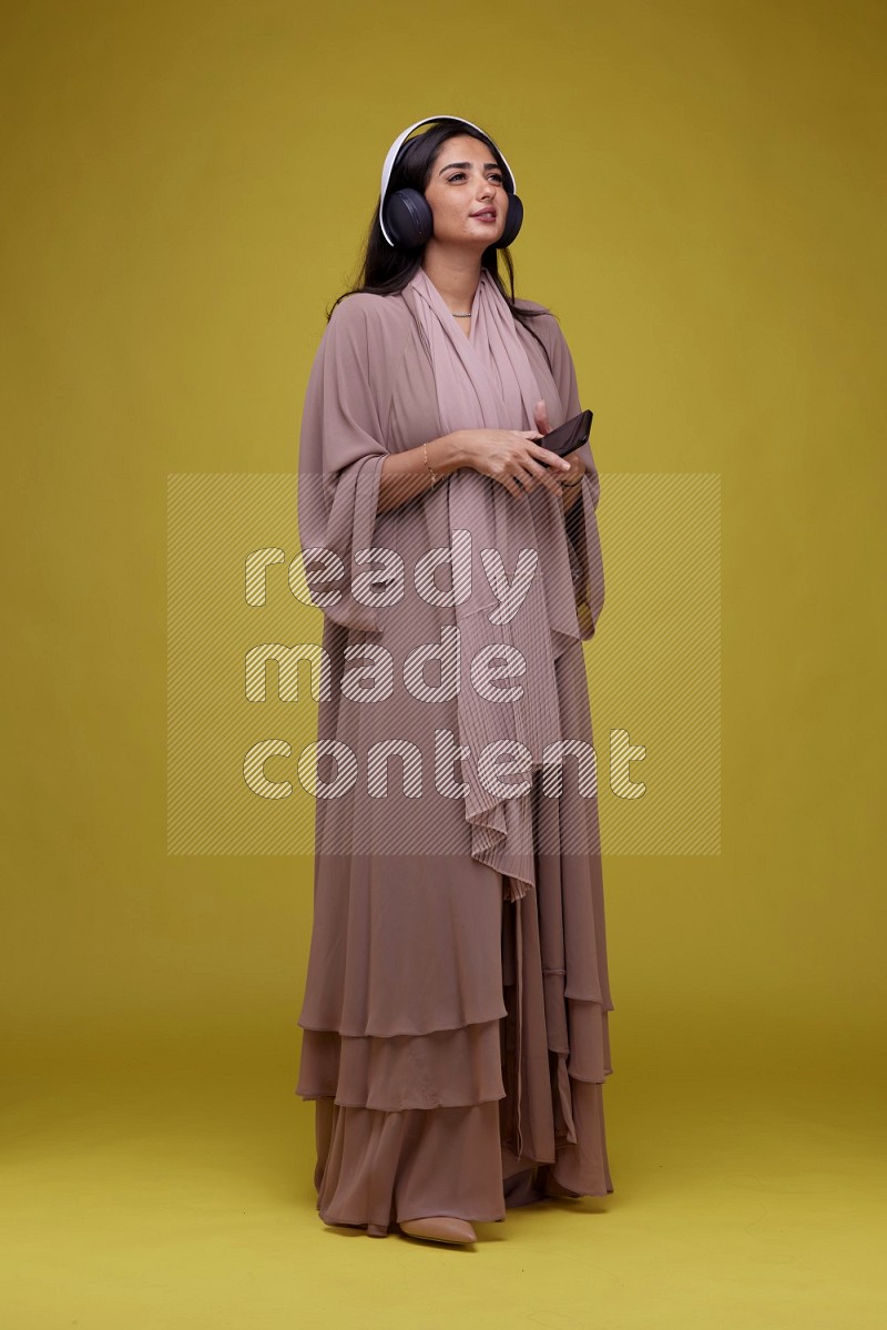 A Saudi woman Listing to Music on a Yellow Background wearing Brown Abaya