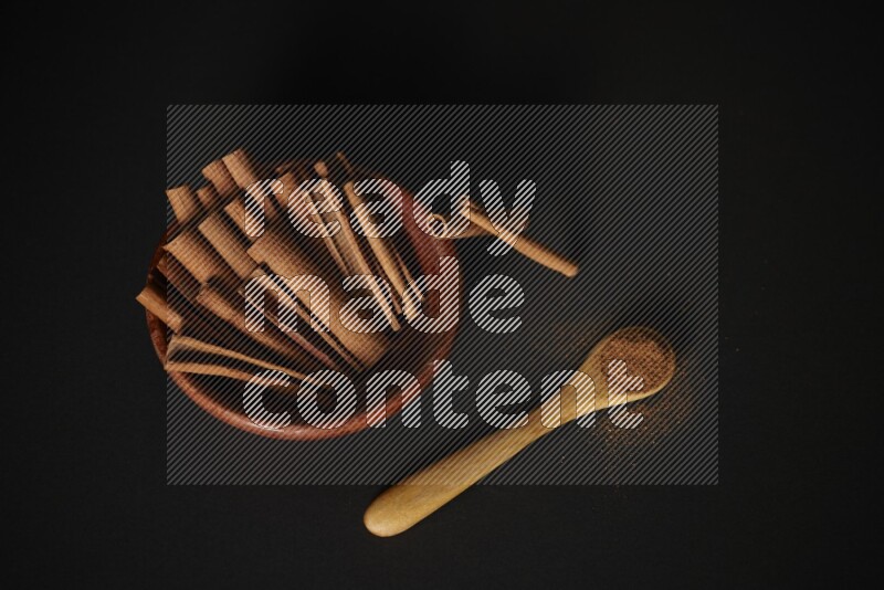 Cinnamon sticks in wooden bowl and cinnamon powder in a wooden spoon on black background