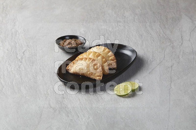 Three fried sambosas in an oval shaped black plate and a sauce in a black round ramekin with two lemon slices on the side on a gray background