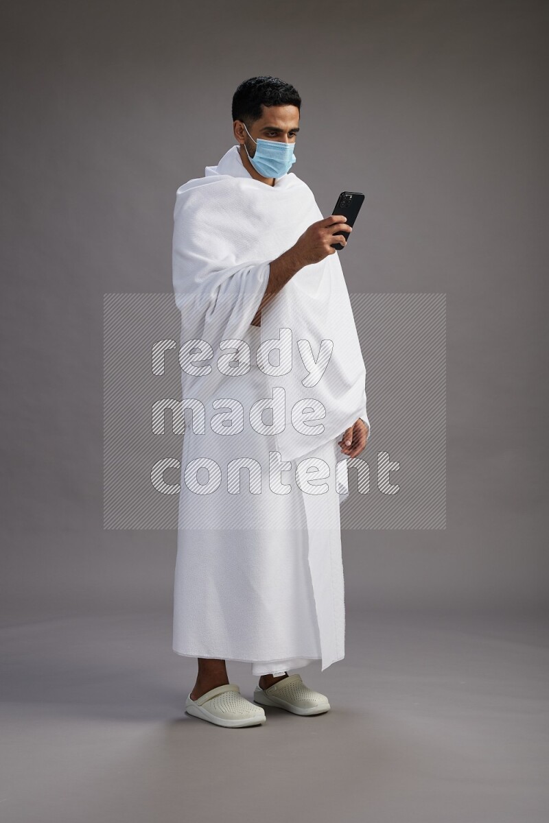 A man wearing Ehram with face mask Standing texting on phone on gray background