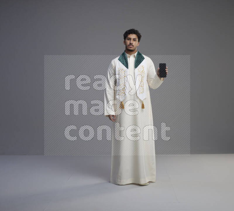A Saudi man standing wearing thob and Saudi flag scarf showing phone to camera on gray background