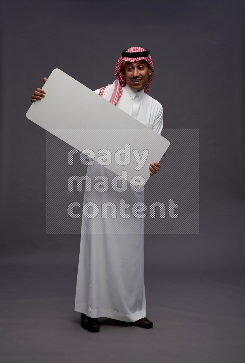 Saudi man wearing thob and shomag standing holding board on gray background