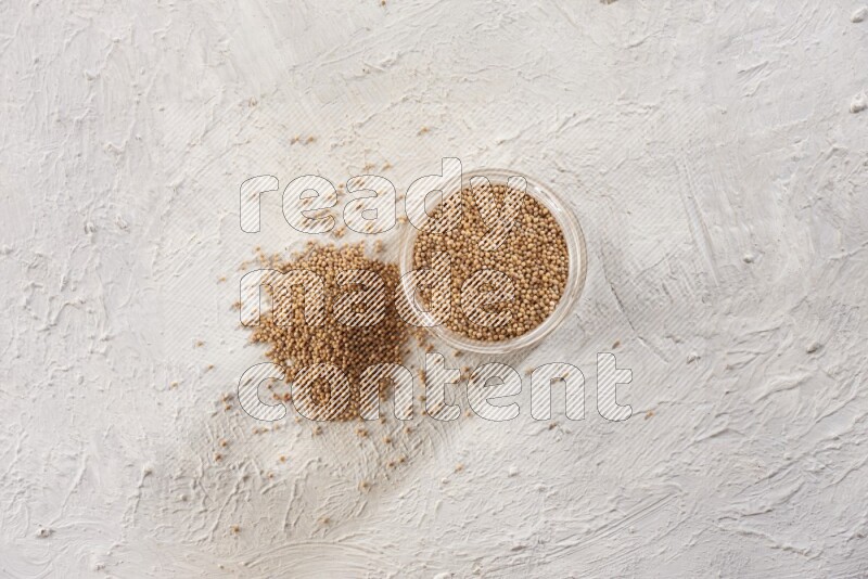 A glass jar full of mustard seeds and more seeds spread on a textured white flooring