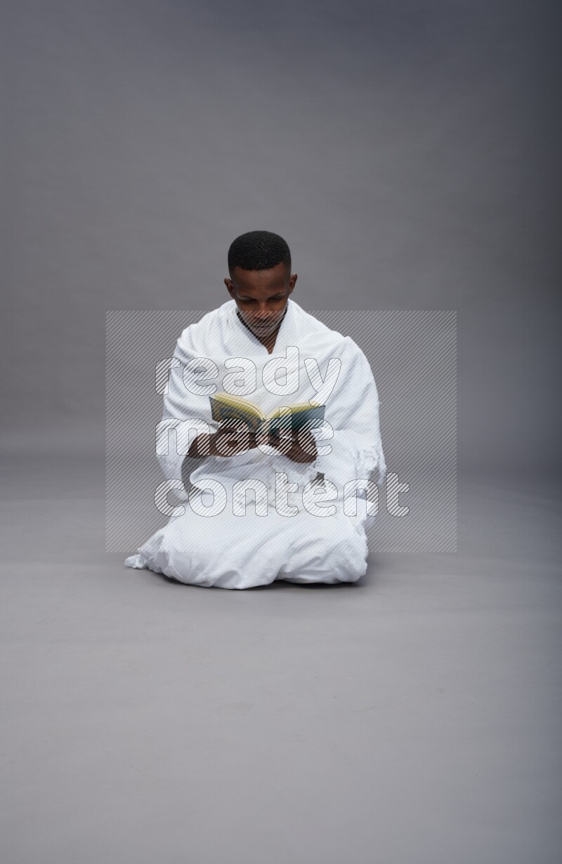 A man wearing Ehram sitting on floor reading quran on gray background