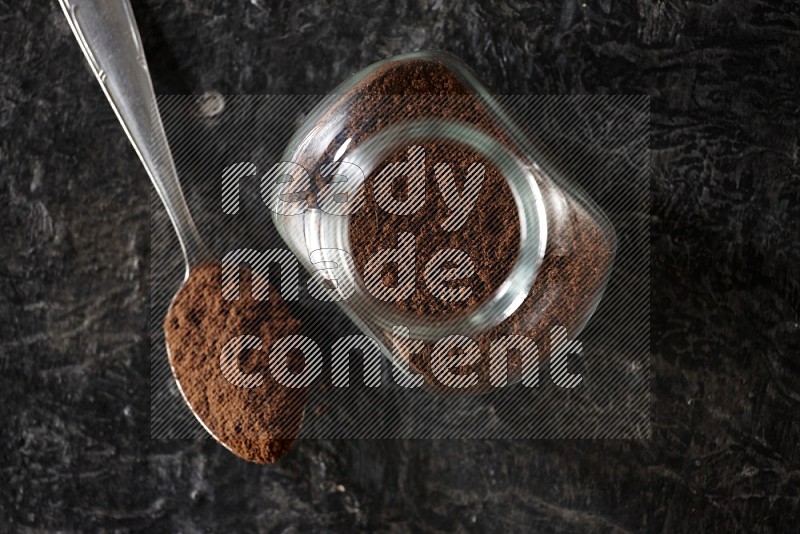 A glass spice jar and a metal spoon full of cloves powder on textured black flooring