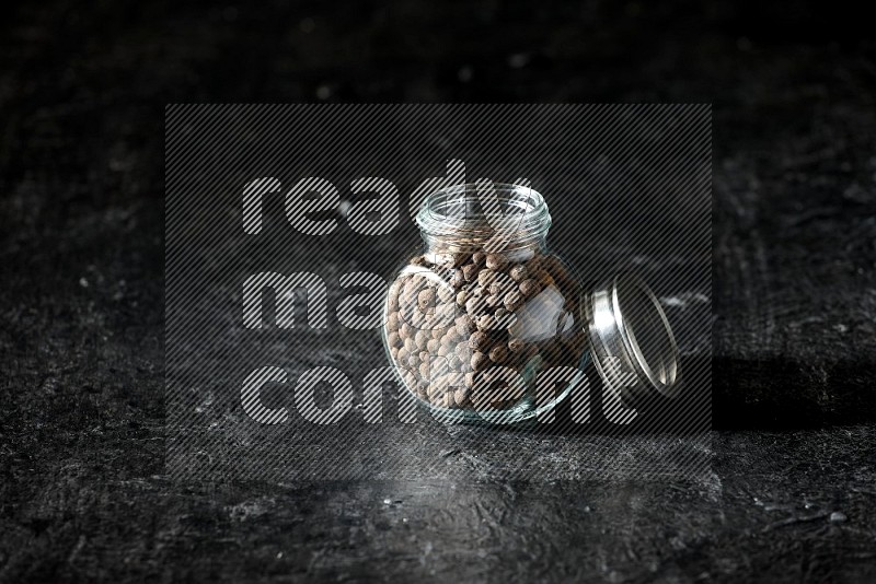 A glass spice jar full of allspice whole balls on a textured black flooring